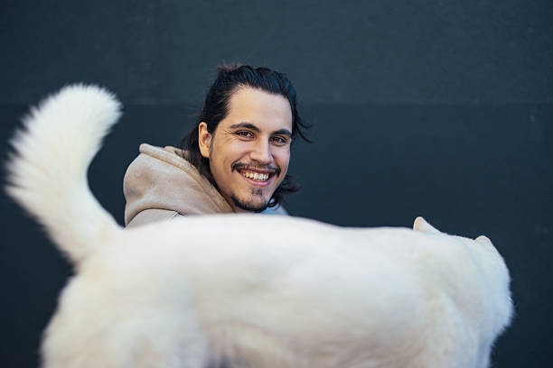 Happy young man sitting on the floor, smiling at his dog whose tail is wagging in front of a simple dark background.