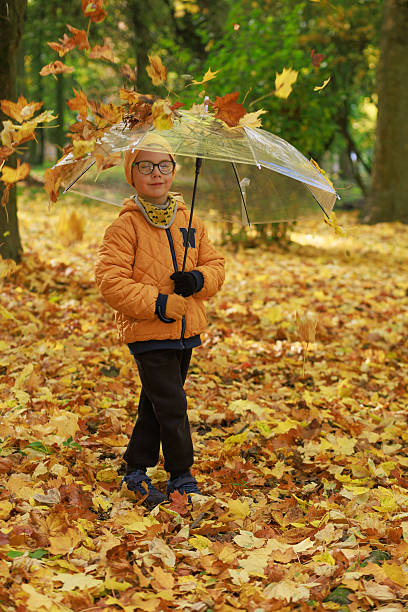 Boy in yellow jacket and hat holds transparent umbrella surrounded by falling leaves.Concept of autumn fashion, childhood and cozy seasonal mood.