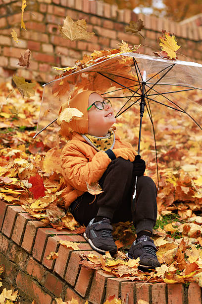 Smiling boy sits under transparent umbrella covered with colorful maple leaves.Concept of joy, childhood wonder and autumn atmosphere.