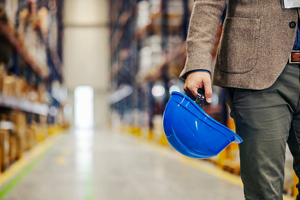 Man in a smart casual outfit holding a blue hard hat in a modern warehouse, symbolizing safety and management