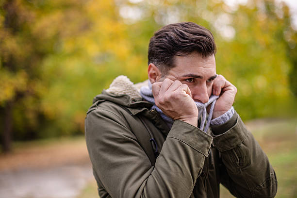 Two adults experience cold symptoms like coughing and sneezing while bundled up outdoors in a park. The fall scenery adds to their discomfort as they deal with illness.