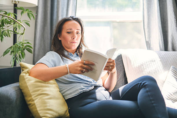 istock woman sitting and reading book on a sofa at home 2243628115