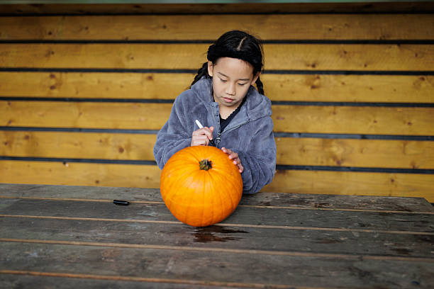 A close-up of a young girl sketching a face on a pumpkin in preparation for carving.