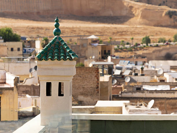 Semi-arid, desert landscape with the Marinid Tombs and structures in the background, a small, white tower-like chimney with a green roof and a decorative finial on top, rises above the rooftop corner of a building in Fez, Morocco., a UNESCO World Heritage site.