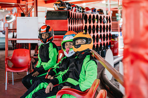 Three friends wearing racing suits and helmets, sitting, anticipating their turn on the go-kart track, seeking excitement