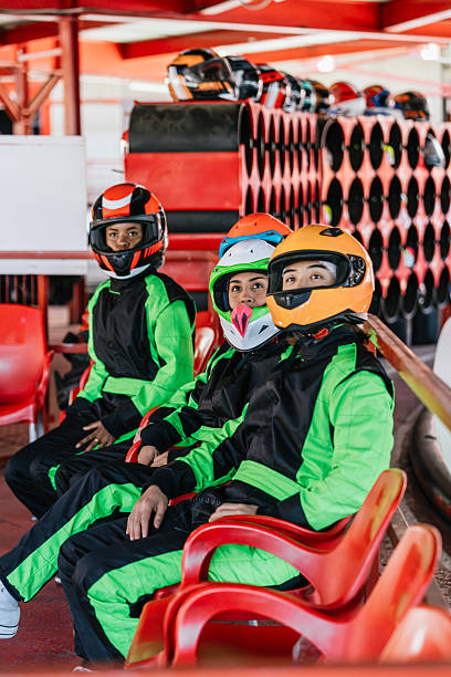 Three young men wearing colorful helmets and racing suits sitting on red chairs, waiting to enjoy go-kart racing