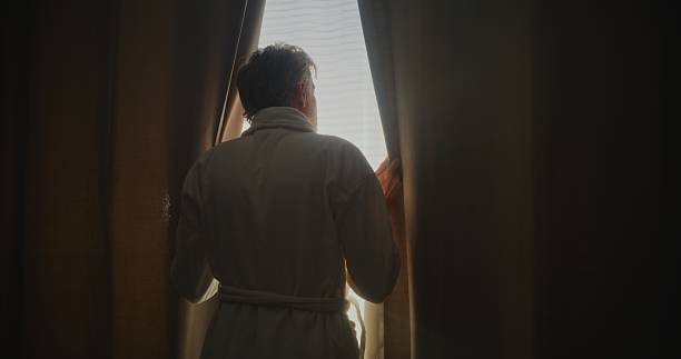 Silver Haired Man Enjoys Soft Sunlight in Stylish Hotel Room. Man Looks Out Window, Pulls Back Heavy Curtains, and Stretches. Concept of Relaxation, While Traveling, Emotional Well Being. Back View.