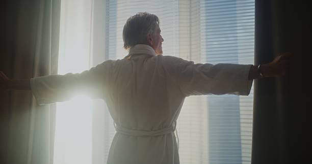 istock Mature Man Looks Out Window, Pulls Back Heavy Curtains, and Stretches 2243663363