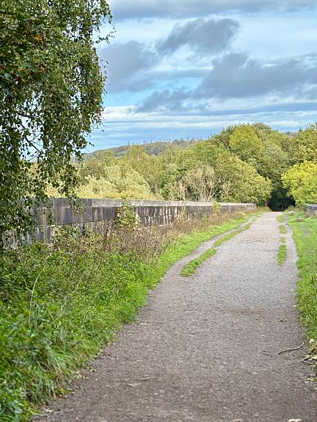 istock A quiet countryside path bordered by stone walls and tall grass stretches into the distance, surrounded by lush green trees and open fields beneath a partly cloudy sky, evoking peaceful calm. 2243665118