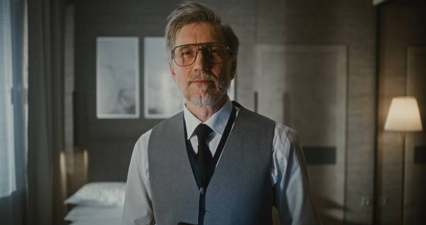 Close Up Gray Haired Man in Vest and Tie Stands, Looking Down at Digital Tablet or Thin Laptop Holds in Hands. Remote Management, Digital Professionalism, Mobile Technology, Business Travel. Portrait.