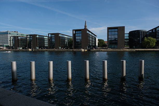 A waterside urban scene showing modern buildings, vertical pier posts, and reflections on calm water. Ideal for real estate marketing, city lifestyle, modern architecture, and travel imagery.