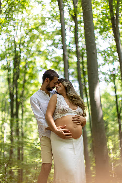 Beautiful young expecting couple is hugging in a forest. Their hands are on her belly. They look peaceful and happy.