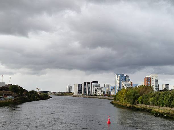 City skyline along a river under a moody sky, Glasgow, Scotland. A wide river, River Clyde flows between two green riverbanks, leading towards a city skyline