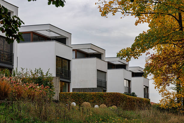 A row of modern houses with gray roofs and white walls are visible in a landscape with autumn foliage.