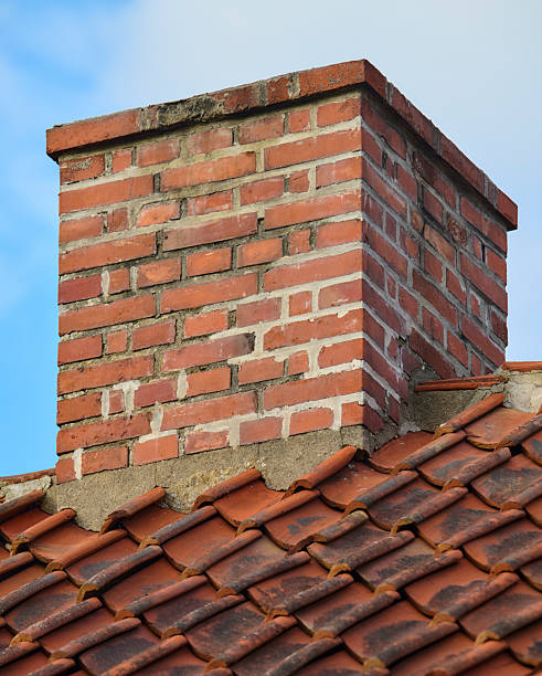 A close-up of a brick chimney rising from a tiled roof, with weathered mortar and rustic textures. Bright blue sky provides contrast to warm red bricks and earthy roof tiles, evoking traditional architecture, home exterior charm, and durable construction suitable for Danish setting visuals.