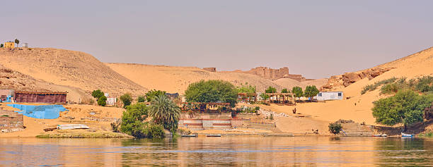 View of the Nile river with sand dunes and lush green areas along the shores in Egypt. The scene features traditional rural buildings, emphasizing the riverâs importance to the ancient landscape.