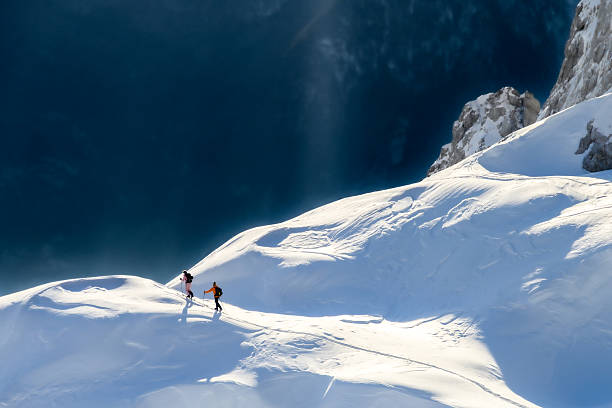 Two climbers ascend a snowy mountain slope under a dramatic, cloud-filled sky. The vast, pristine snowfield and rugged peaks highlight the adventurous spirit and challenge of winter mountain climbing. The scene captures the essence of exploration and endurance in a breathtaking, remote landscape.