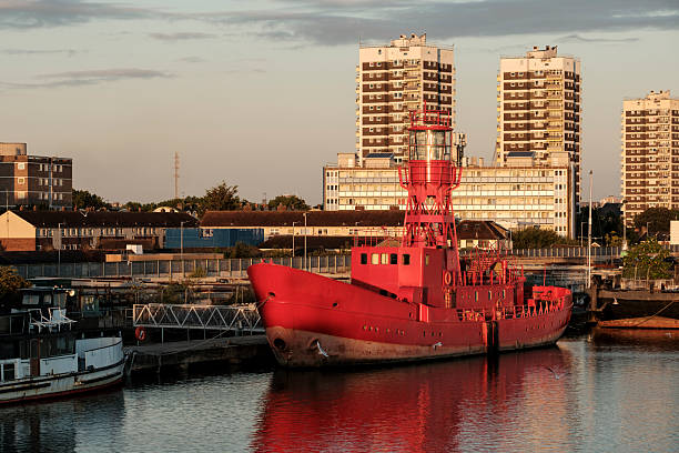 Red ship with a tower structure moored in a bustling urban harbor, reflecting on calm water with apartment buildings towering in the background under soft golden light