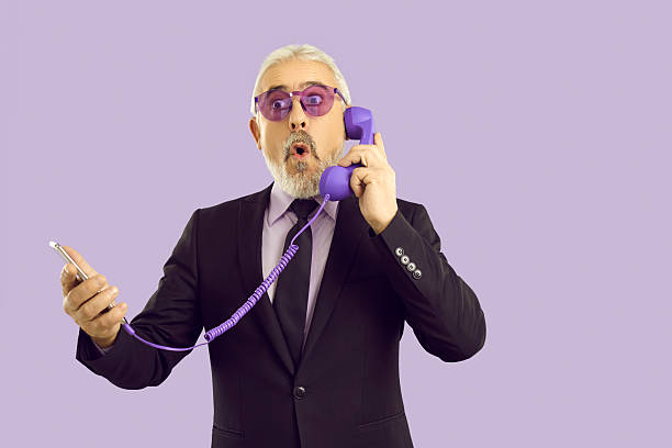 Stunned senior businessman in suit isolated on purple studio background shocked by news on cellphone and landline handset. Surprised mature man talk on smartphone with funny set.