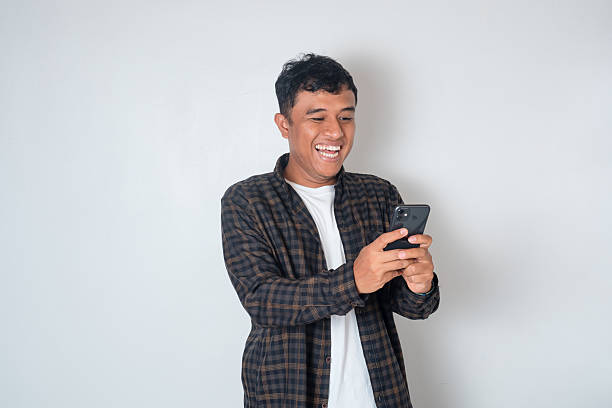 istock Asian man smiling while holding smartphone with both hands, casual and cheerful expression. Concept of communication, chatting, social media, and positive lifestyle on white background. 2243720807