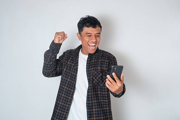 istock Asian man celebrating success while looking at smartphone, expressing happiness and excitement. Concept of achievement, good news, winning, and joyful reaction on white background. 2243720872