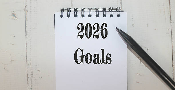 istock Hand Writing 2026 Goals on Notebook with a clock, glasses, and planner on the desk. New year planning, motivation, productivity, and goal setting for 2026. 2243722666