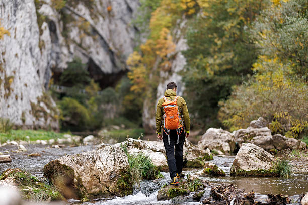 Rear view shot of male backpacker exploring majestic autumn landscape with mountain river among rock formations