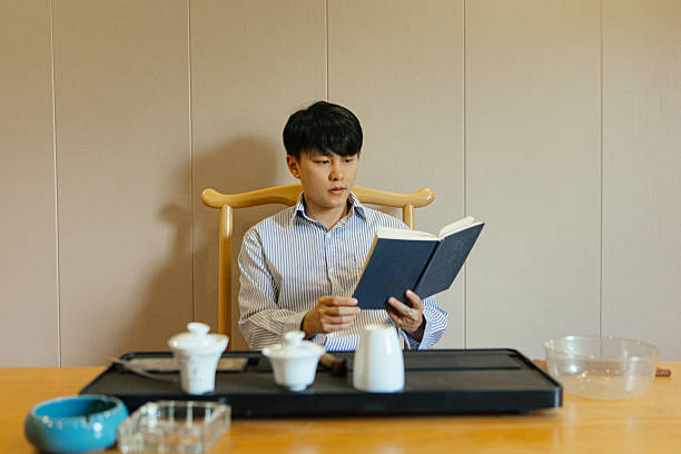 Young Asian man sitting at a tea table drinking tea and reading a book in a calm indoor setting, symbolizing relaxation, mindfulness, and a peaceful lifestyle inspired by traditional Asian culture and modern simplicity.