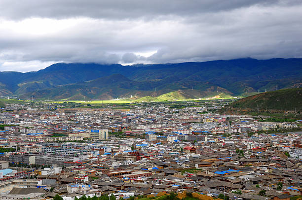 A panoramic view showcases a town nestled among mountains with a cloudy sky overhead, featuring various buildings and urban layout.