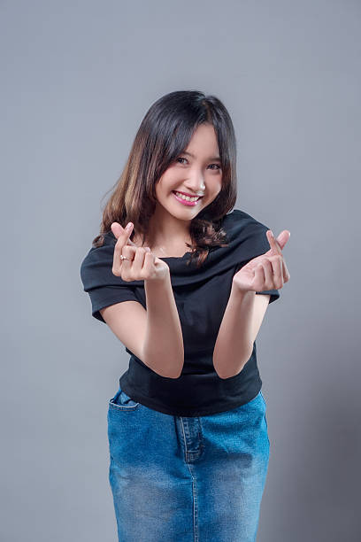 istock A cheerful young Asian woman smiling brightly while making a finger heart gesture with both hands, expressing love, friendliness, and positive energy in a casual modern appearance. 2243735214