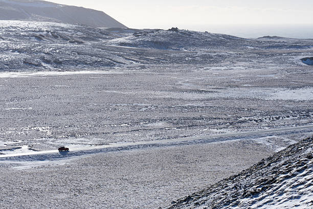 Red vehicle driving on a long road winding through a remote, snow-dusted icelandic landscape
