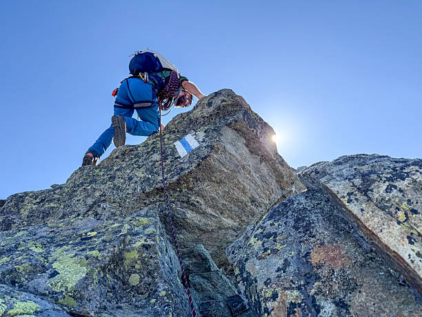 Man climbing steep rock face with safety rope and gear under clear blue sky. Outdoor mountaineer scaling rugged mountain terrain. Adventure sports and alpine climbing activity.