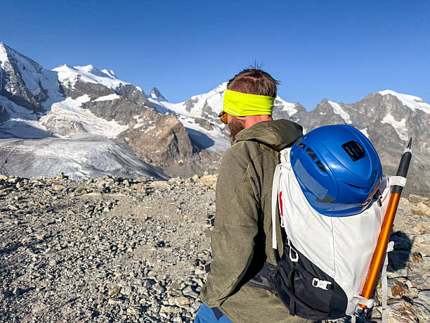 Man with backpack and climbing helmet hiking on rocky terrain near snow-covered peaks. Outdoor adventure in alpine mountains with clear blue sky. Mountain trekking and nature exploration.