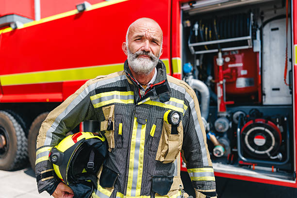 Mature Caucasian firefighter in full uniform standing confidently beside a bright red fire engine. The scene is set outdoors under clear daylight, showcasing the firefighter's readiness and determination.