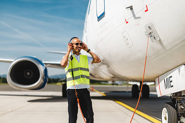 Mid adult Hispanic male wearing a high-visibility vest, standing beside a cargo aircraft, adjusting a headset while performing a systems check on a sunny day.