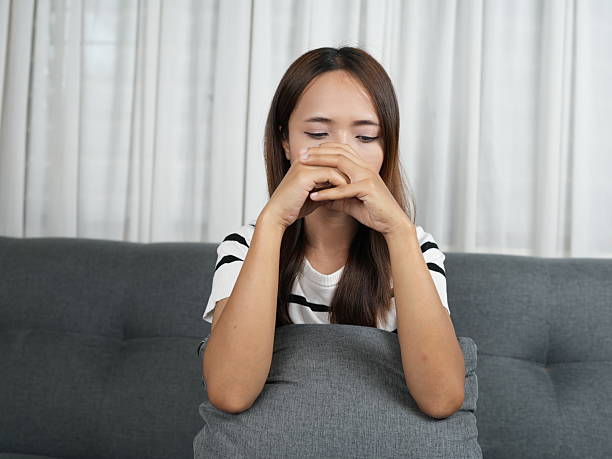 istock Young woman expressing contemplation, sadness, or anxiety, resting hands on face while sitting on gray couch in a cozy indoor setting with natural light 2243747971