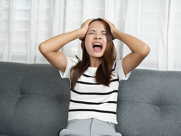istock Young woman expresses frustration while sitting on gray sofa, holding her head with both hands. She wears striped shirt and appears overwhelmed by emotions 2243748176