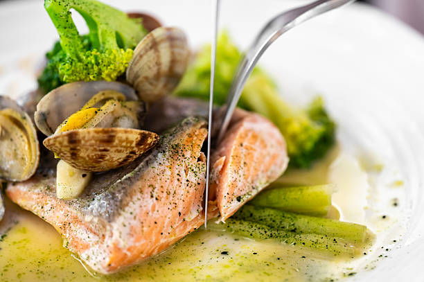 A fork cuts into a beautifully plated fish fillet, served with clams and green vegetables in a savory sauce on a white plate.