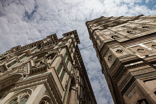 Low angle view of Giotto’s Bell Tower and adjacent cathedral facade in Florence featuring intricate Gothic architecture against a cloudy sky. The detailed stonework and towering structures highlight the historical significance of this iconic Italian landmark.