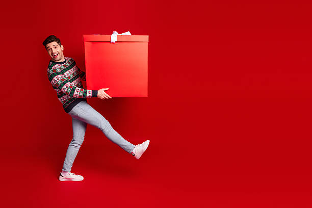 istock Funny man in christmas sweater carries oversized gift box against a bold red backdrop creating a playful holiday moment 2243754499