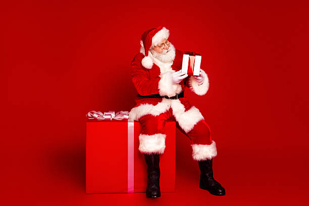 istock Santa Claus sits on a red gift box holding two presents with a white beard and red suit against a solid red background ready for Christmas sale and seasonal celebration 2243757808