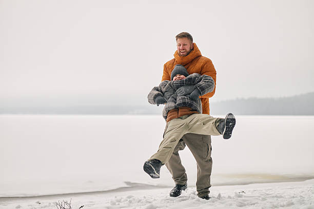 Caucasian man spinning child in winter clothing outdoors on snowy landscape near frozen lake, both smiling and enjoying playful moment together