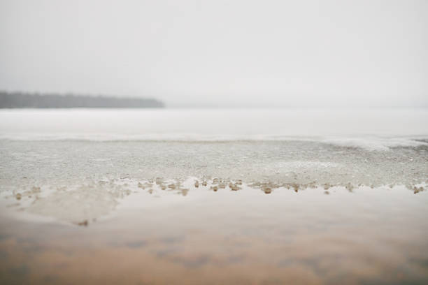 Frozen lake surface melting near shoreline with thin layer of ice and water reflecting overcast sky, distant treeline visible in background, winter landscape showing seasonal transition