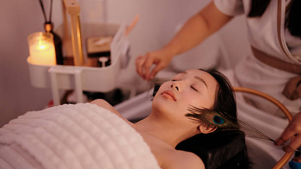 Asian woman relaxing as a spa therapist gently stimulates her face with feathers during a tactile therapy session.