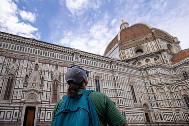 Young adult traveler wearing a blue backpack exploring Florence Cathedral and Brunelleschi’s Dome. The detailed architecture and partly cloudy sky create a stunning backdrop. The traveler gazes upward, appreciating the historical structure in Florence, Italy.