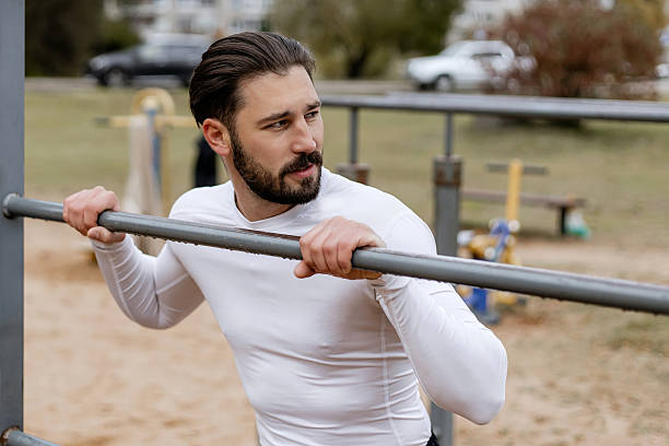 istock Close-up of a young bearded man in a white compression shirt doing pull-up exercise on outdoor fitness bars, focusing on upper body strength and determination in a park setting 2243769654