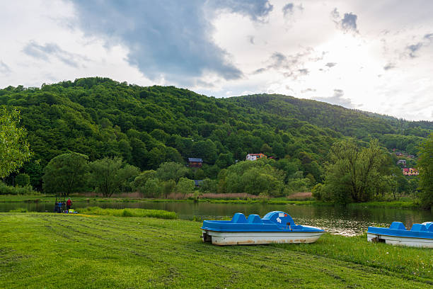 Porabka, Poland – May 24, 2025: Blue pedal boats rest on the grassy shore of a small lake surrounded by wooded hills and village houses. The quiet setting evokes rural leisure and summer tourism.