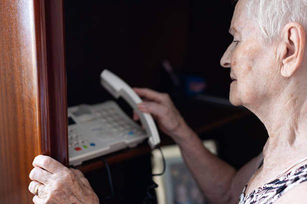 Mature white-haired woman in profile holding a landline receiver to her ear, dialing to connect with family or friends at home, conveying everyday communication and care