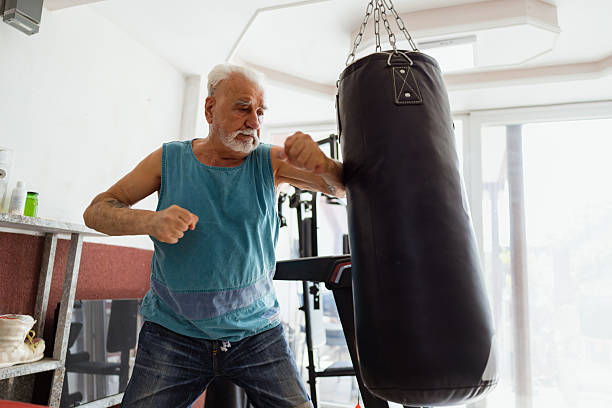 An older man trains with a heavy punching bag in a bright gym, displaying strength, focus, and determination during a fitness workout.