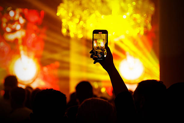 Crowd at a concert with a hand holding a smartphone capturing performance under bright yellow stage lighting.